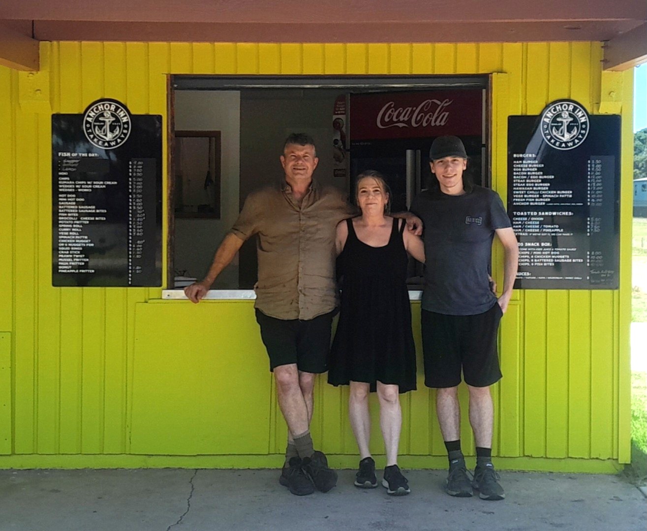 A family of three stands smiling in front of a bright yellow food stand with a service window. Menus are on either side.