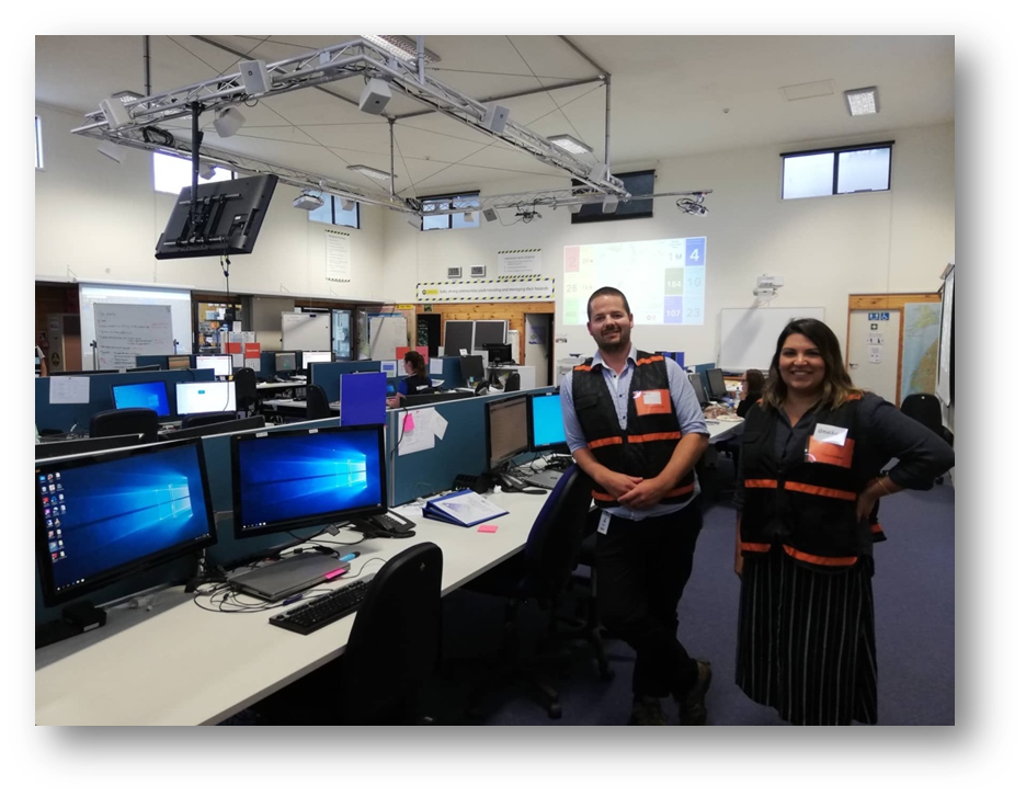 two staff stand in a civil defence control centre with monitors and computers all around them.