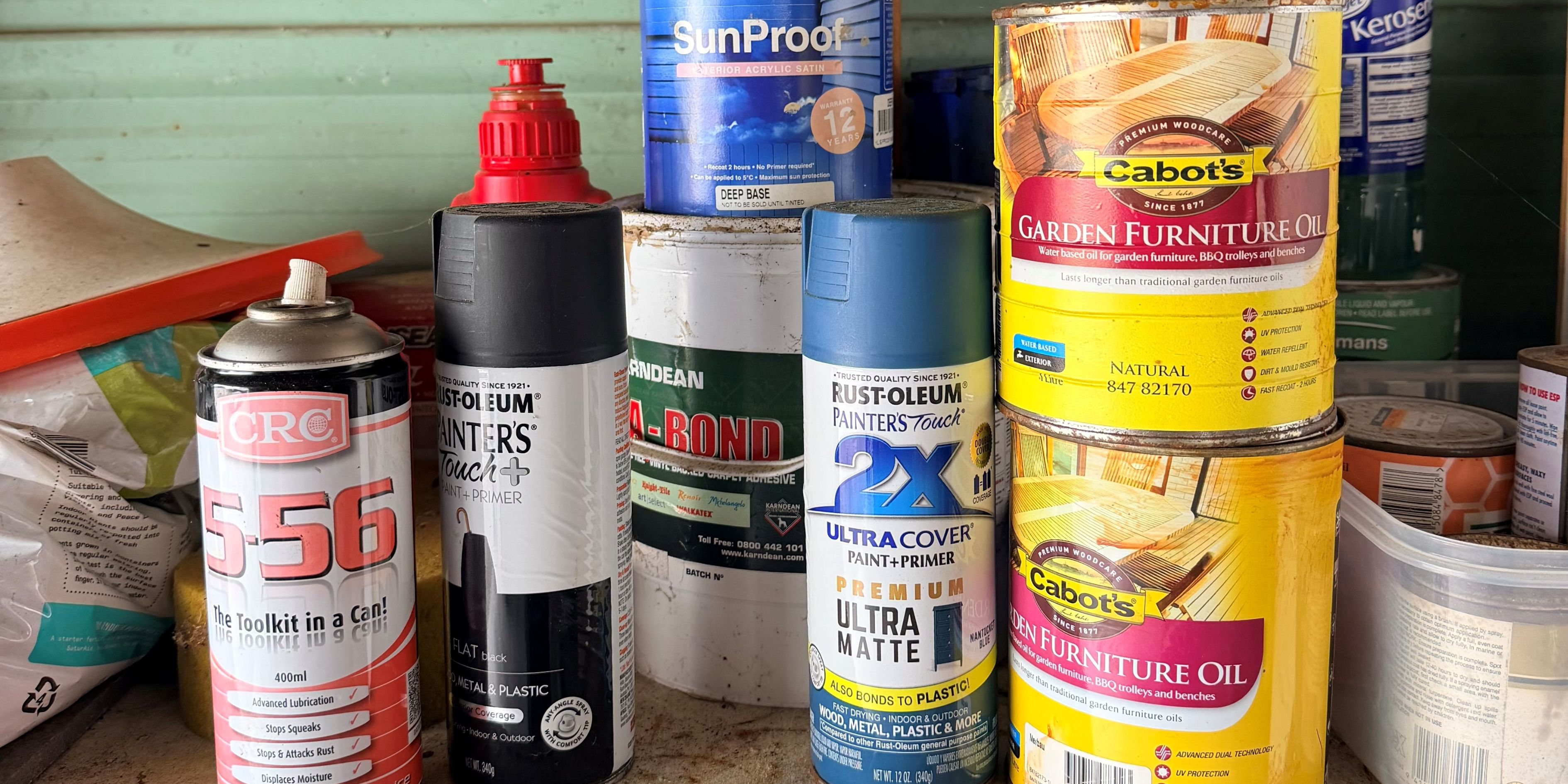 A cluttered shelf with various paint cans and spray bottles, including brands like Taubmans and Cabot's. Background is a green wooden wall.
