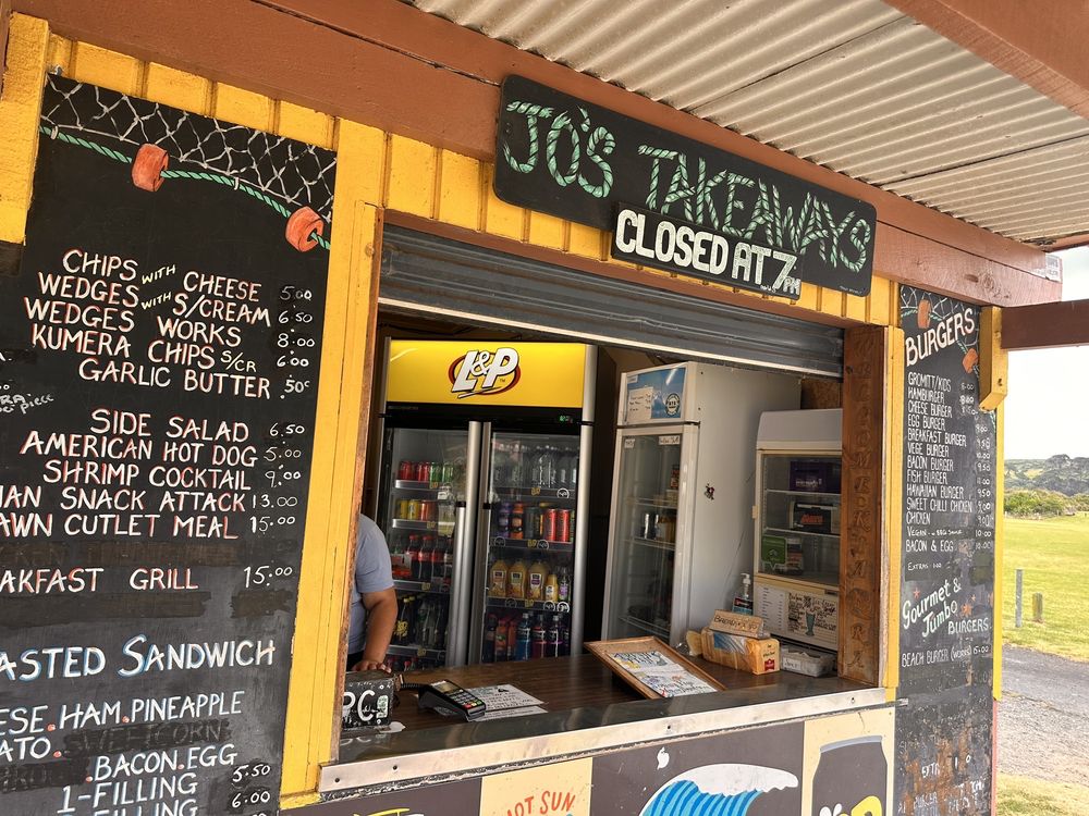 Outdoor takeaway stand with chalkboard menus listing various foods, like burgers and chips. A fridge with drinks is visible inside. Sign says "Jo's Takeaway, Closed at 7." Casual, inviting vibe.
