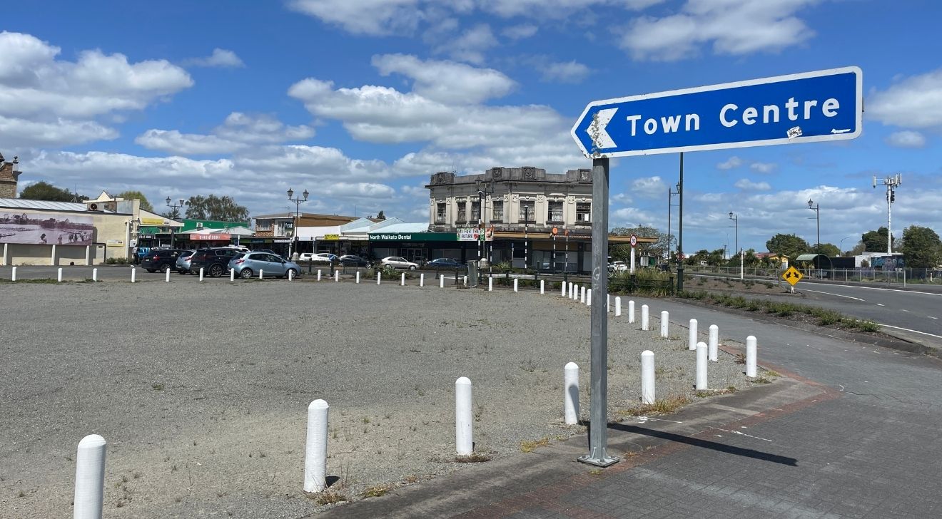 Blue sky with scattered clouds over a town intersection. A sign reads "Town Centre" pointing left. Cars parked around historic building in the background.