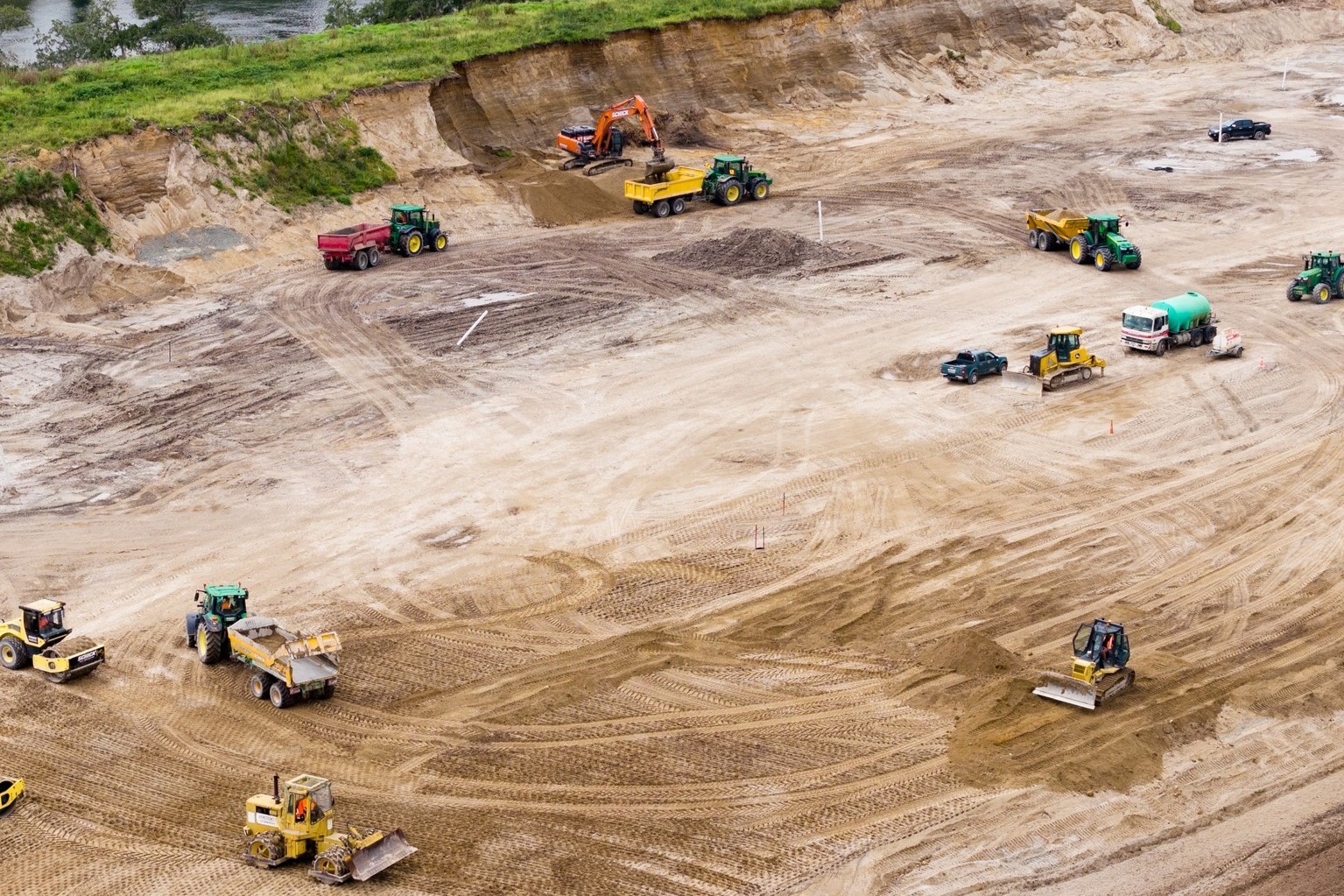 Aerial view of a large construction site with various equipment, including excavators, bulldozers, and trucks. The scene is active and industrious.